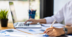 Close-up image of a businessperson working on a laptop and reviewing charts on a desk.